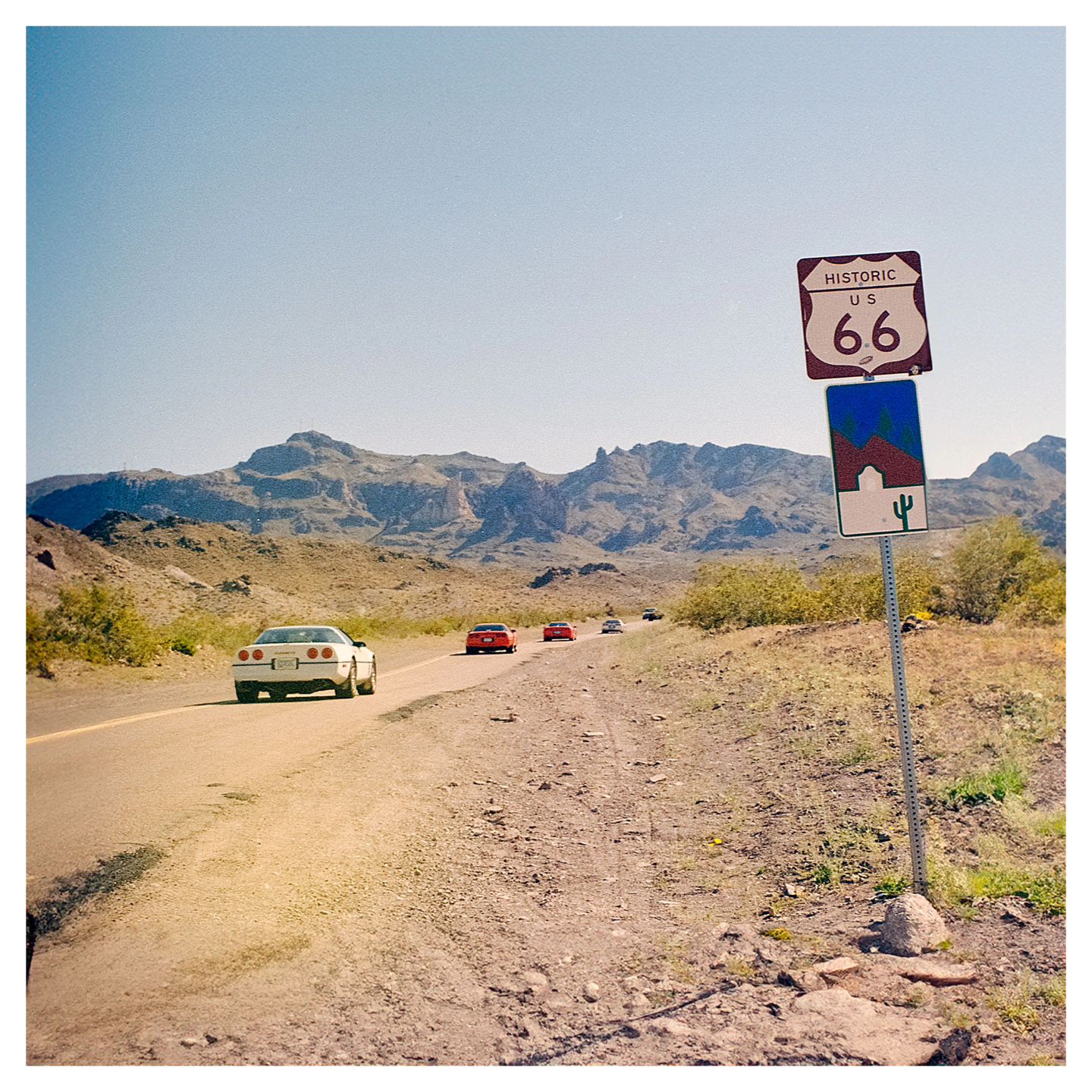 The Route 66 sign atop a lonely pole on a stretch of desert road with mountains in the background a four cars driving towards them.